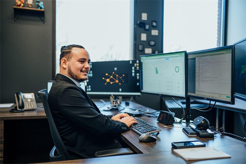 Jesus Sosa, founder of Express Automation Solutions, smiling at his desk in front of three monitors showing dashboards and code